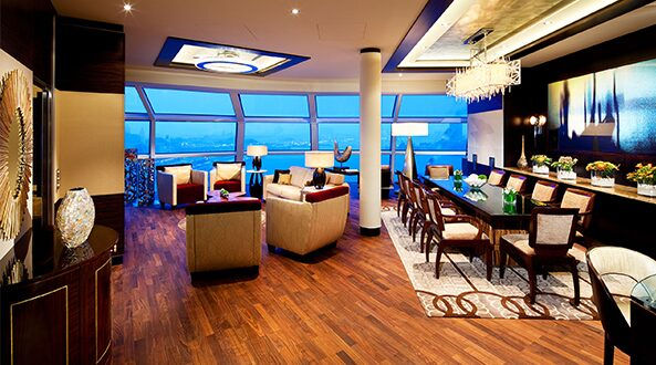 Luxurious room with modern furniture, large dining table, and vibrant decor. Floor-to-ceiling windows show a serene blue sky. Warm lighting.