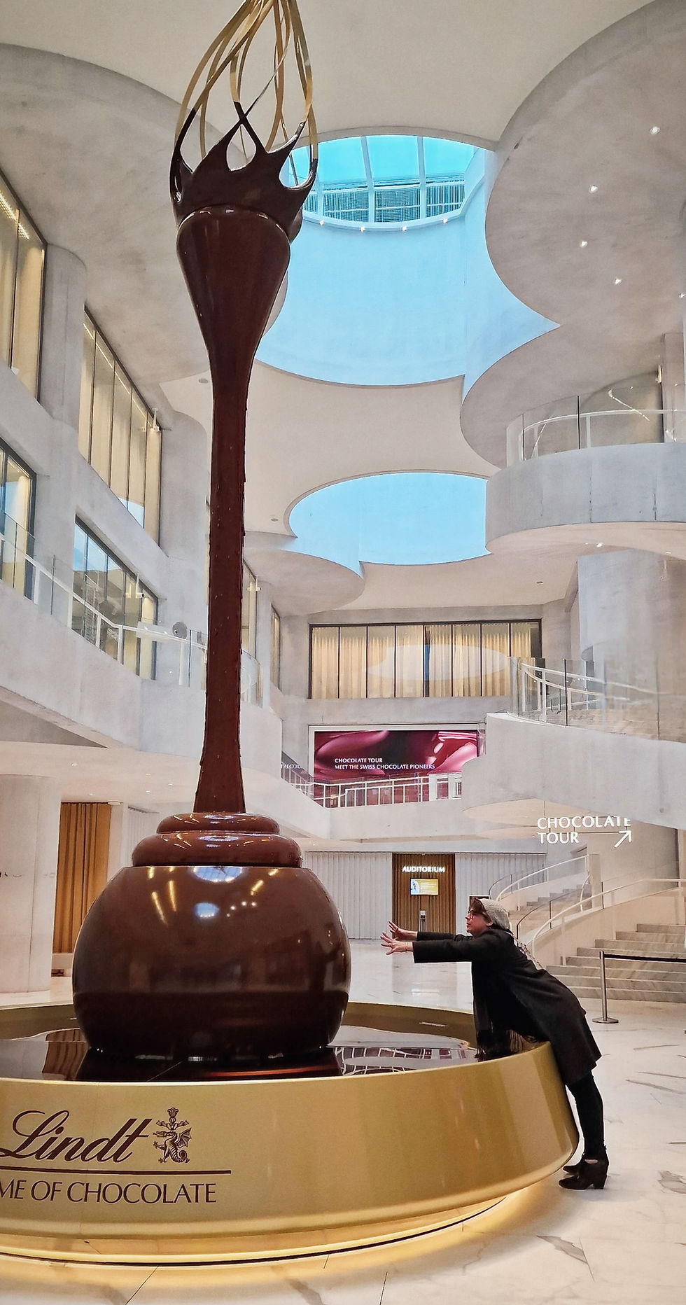 A person poses near a large chocolate display at Lindt Home of Chocolate, with modern architecture in the background. Text: Chocolate Tour.