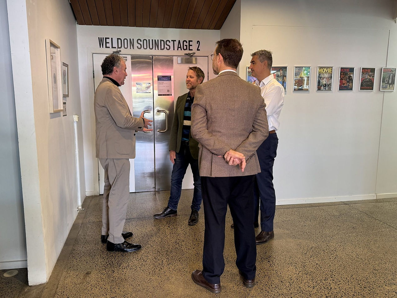 A group being shown the entrance to the Weldon Soundstage at Chrysaor Studios, with framed film magazine covers displayed along the wall.