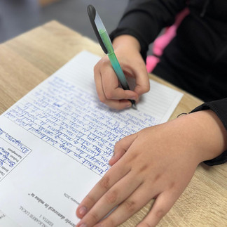 Detaliu cu mâna unui elev care scrie cu atenție pe o foaie de hârtie liniată.