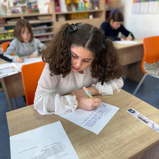 O elevă cu părul creț, concentrată, scriind la masa de lucru în sala de clasă.