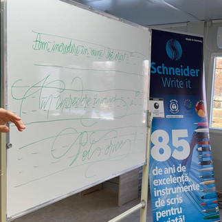 O tablă albă pe care sunt scrise mesaje motivaționale în limba română, lângă un banner publicitar pentru instrumente de scris.