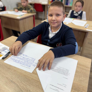 Un elev așezat la bancă, zâmbind spre cameră, având în față o foaie de examen și un pix.