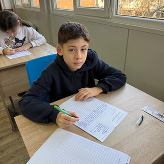 Un elev așezat la birou, privind spre cameră, cu materialele de scris pregătite pe bancă.