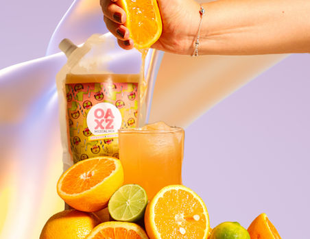 The citrus-flavored punch is showcased, a perfect combination of orange and lime, creating a succulent citrus flavor that blends with the mezcal, resulting in an explosion of taste. The graphic depicts a woman's hand squeezing half an orange over a tall glass of the prepared drink on the rocks, surrounded by halved oranges and limes. The punch is arranged to resemble a mountain against a light lilac rainbow background.