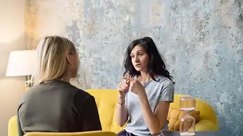 Two women engaged in a therapy session, sitting comfortably and talking in a warm, professional office setting.