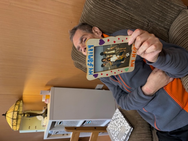 Man in an armchair holding a photo frame labeled "My Family, My Heart." Cozy room with a lamp and shelves, creating a warm atmosphere.