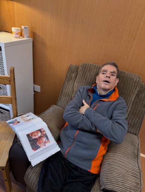 Man sitting in a chair, looking relaxed, with a photo album open on a nearby table. Brown wood wall and furniture in the background.