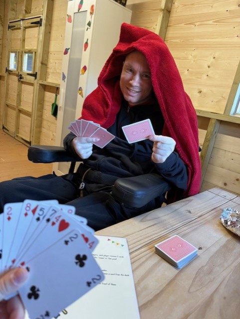 Person in red blanket plays cards in wooden room, smiling. Hand with cards in foreground, wooden table with deck visible. Cozy mood.