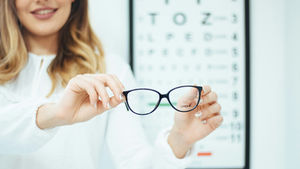 Smiling woman holds black Vogue eyeglasses in front of an eye chart in a bright optician's office.