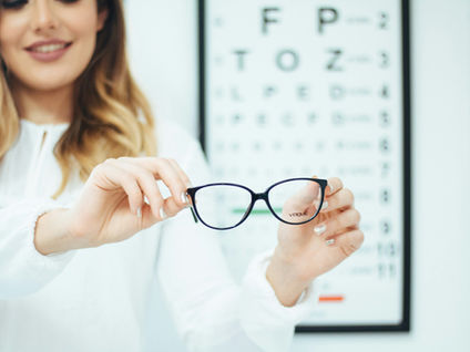 Smiling woman holds black Vogue eyeglasses in front of an eye chart in a bright optician's office.