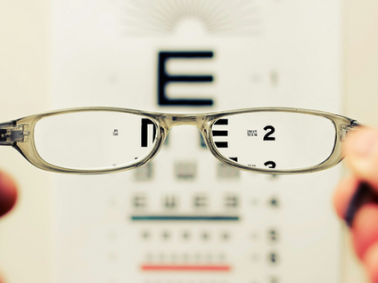 Glasses held in focus over an eye chart, with the letter "E" visible. Blurred background, calm setting, neutral tones.