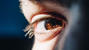 Close-up of a brown eye with long lashes on a face, lit warmly against a dark blue background, creating a thoughtful mood