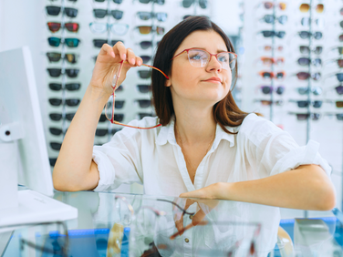 Woman in a white shirt in an eyewear store holds glasses, looking thoughtful. Glasses display in the background. Bright, clean setting.