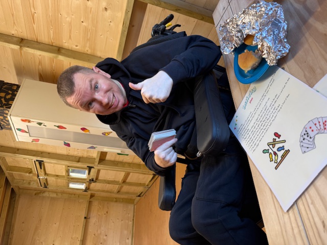 Man in a wheelchair holds cards, smiling with thumbs up in a wooden room. Table has a paper with text and a foil-wrapped snack.
