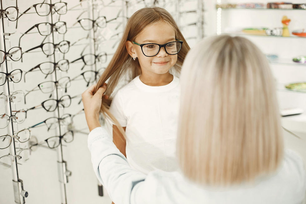 Girl trying on black glasses as an optician adjusts her hair in an eyeglass shop, with rows of frames behind her.