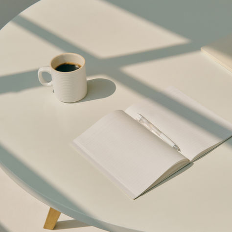 Open notebook and pen on a round table with sunlight casting shadows. A cup of black coffee sits nearby, creating a serene workspace.