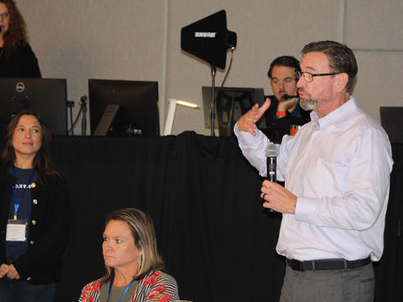 Sales professional speaking into a handheld microphone during a breakout session, sharing real-world sales insights while team members and production staff listen and support the discussion.