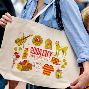 A gold, yellow, and red canvas tote bag reads "Soda City - Brain, Body, Belly - Cola, SC" designed by Rachel Sullivan