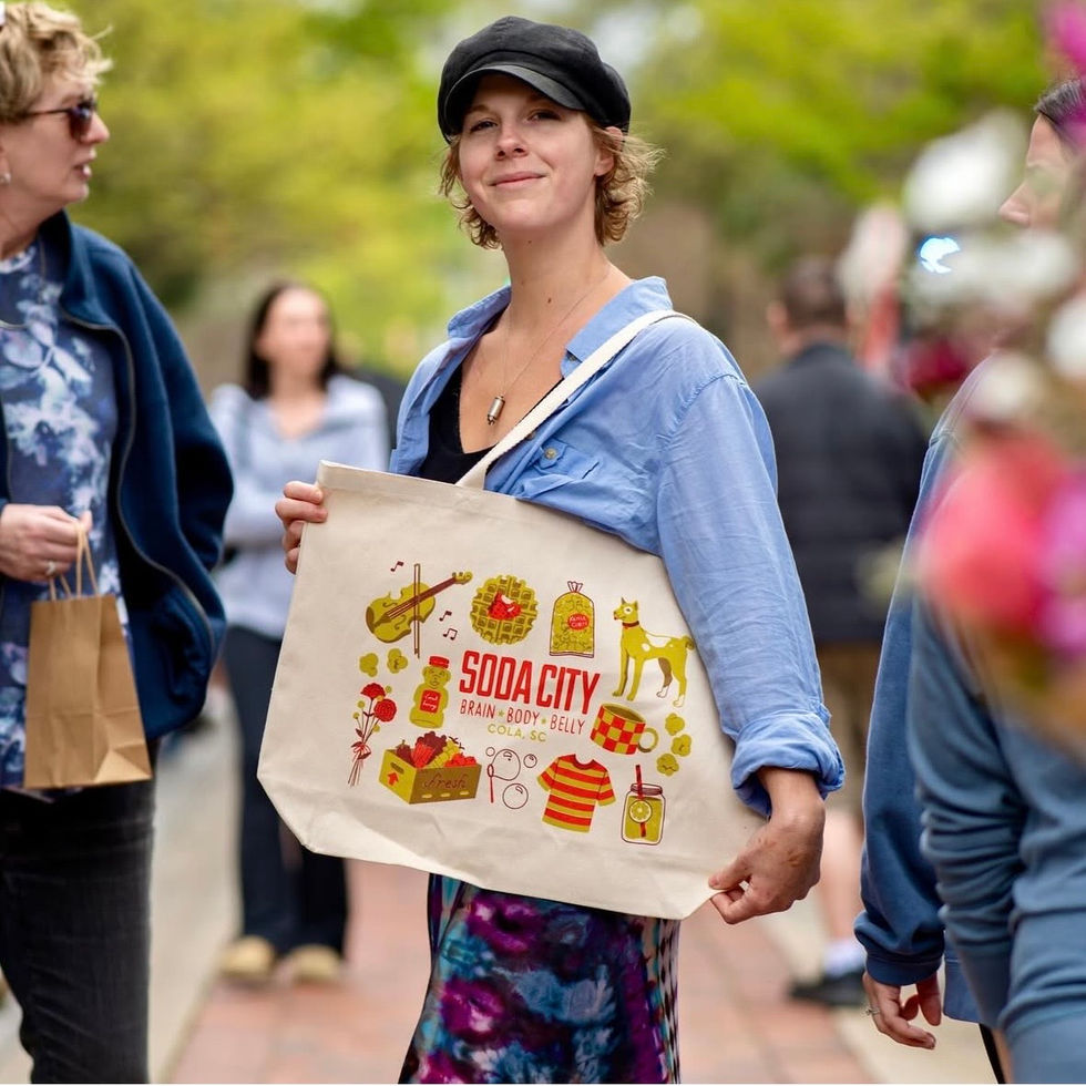 A smiling woman holds a colorful tote bag featuring illustrations of a dog, waffle, popcorn, mug, honey, violin, lemonade