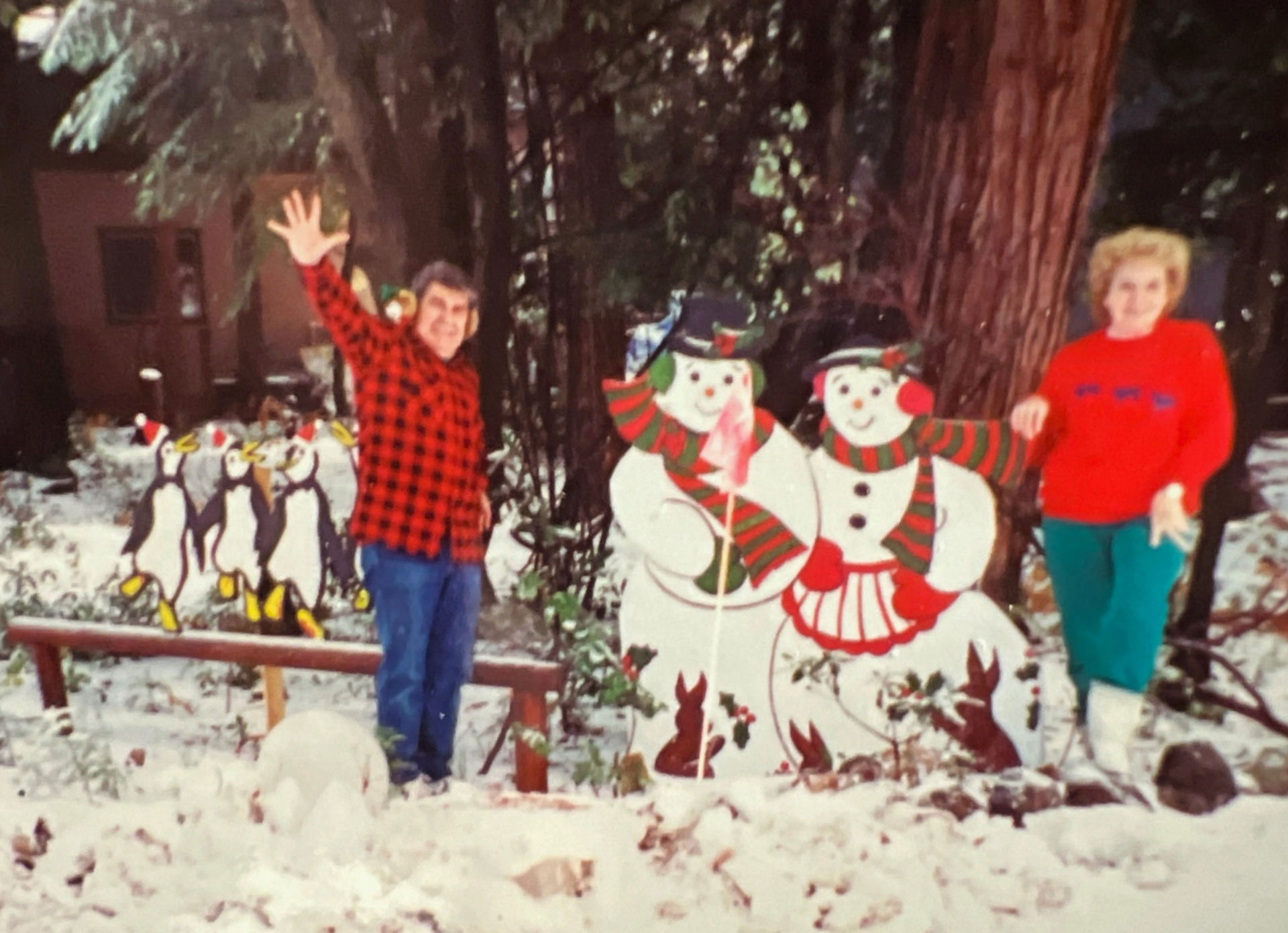 The image shows Leslie Carrara-Rudolph's parents, Art and Doris, in the backyard surrounded by their wood cutout decorations. Art waves to the camera, wearing a red and black flannel button up shirt with blue jeans. Doris wears a red sweatshirt with teal pants. To the left of the screen are three wooden cutouts of penguins dancing wearing red and white Santa hats. To the right of the screen are a wooden cutout of two snowmen with raised eyebrows, smiles, and round, three-dimensional orange noses. They both wear red and green striped scarves and holly on their heads. The snowman on the left of the screen wears green earmuffs and mittens as well as a black top hat. The snowman on the right wears red earmuffs and mittens as well as a red and white striped apron. Below them are two brown bunnies surrounded by holly. Art stands between the penguins and the snowmen while Doris stands all the way to the right.