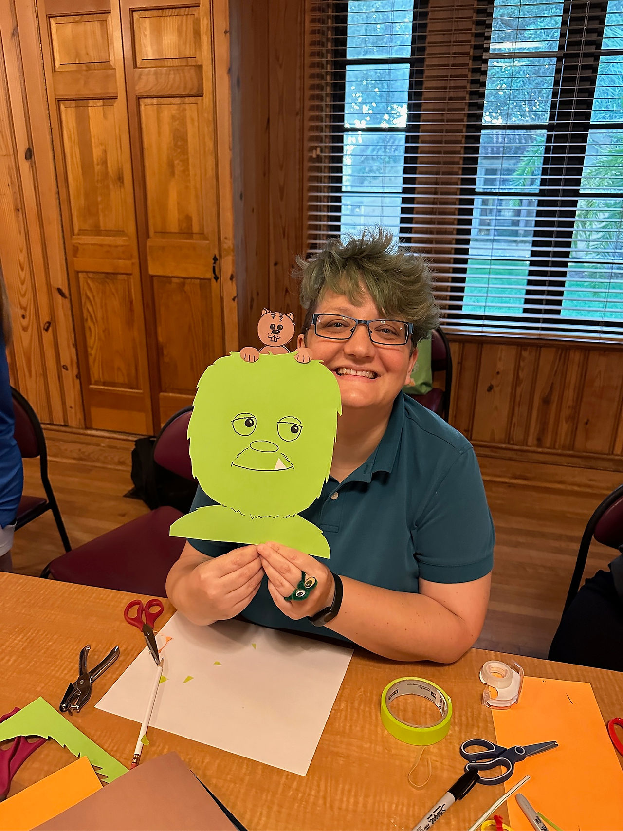 An individual smiles holding their puppet, a green furry creature smiling with a white tooth popping out. On top of the puppet is a brown squirrel-like creature with buck teeth, a tail sticking straight up, and small paws on top of the green creature's head.