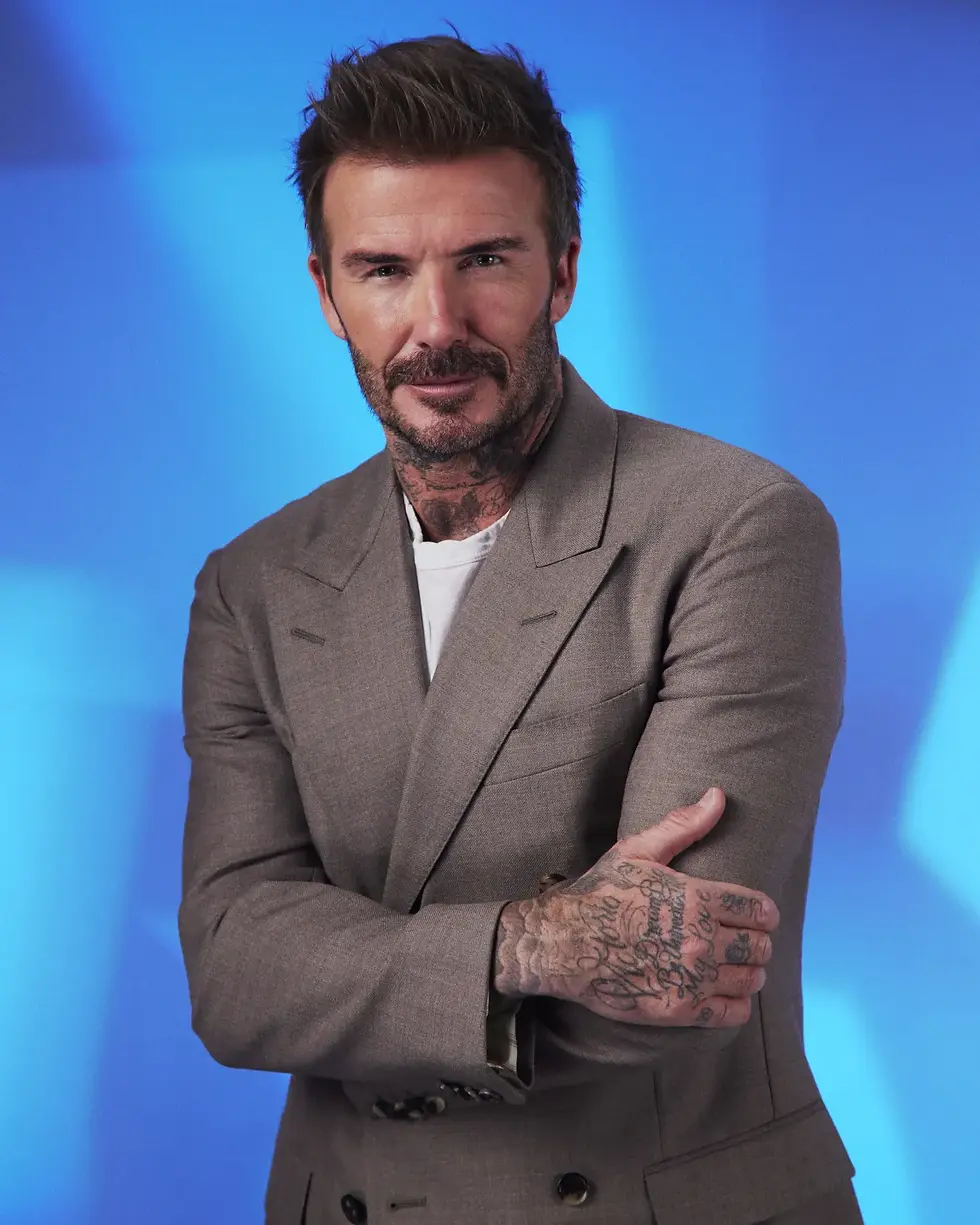 David Beckham in a brown blazer poses with arms crossed against a blue studio background, looking confident.