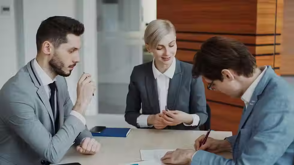 Three professionals in business attire reviewing and signing documents together at a meeting table in a modern office.