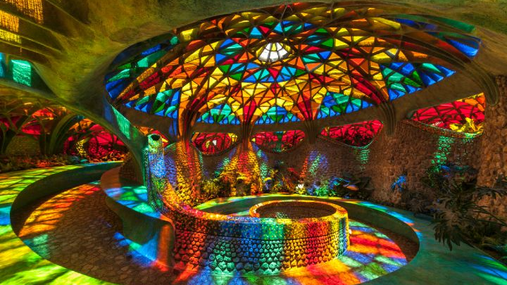 Colorful stained-glass dome interior with curved stone benches and rainbow light patterns across the floor and walls.