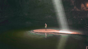 A person stands on a rock platform in a cave, illuminated by a beam of light from above. The water around is clear and turquoise.