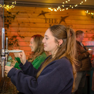 A young woman using a self-service drink tap at an event. She is wearing a dark sweatshirt and has her hair tied back. In the background, other people, including Catherine in a green sweater, are also using the taps and enjoying drinks. The setting has a warm atmosphere with fairy lights and wooden decor.