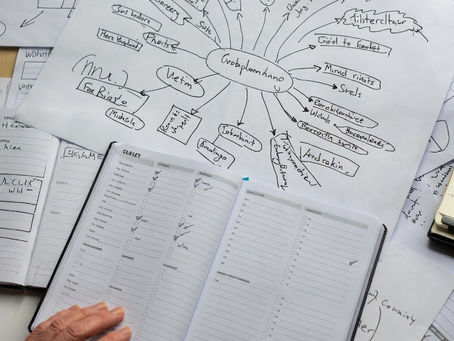 Hands hold an open planner with checklists. Papers with mind maps and notes are scattered on a table. A focused, organized workspace.