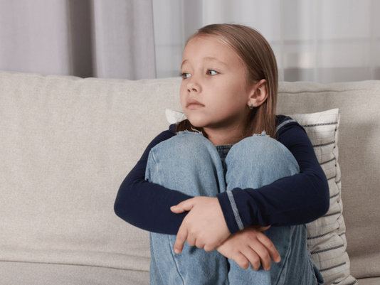 Ein Mädchen sitzt auf der Coach, umarmt seine Knie und schaut mit traurigem Blick weg.