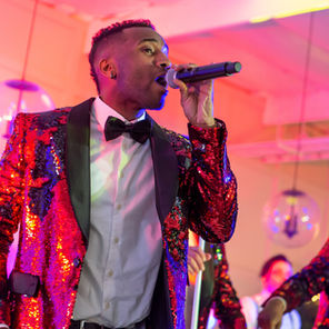 Male singer in a red sequined tuxedo sings into a microphone on a pink-lit stage with band members blurred behind him.