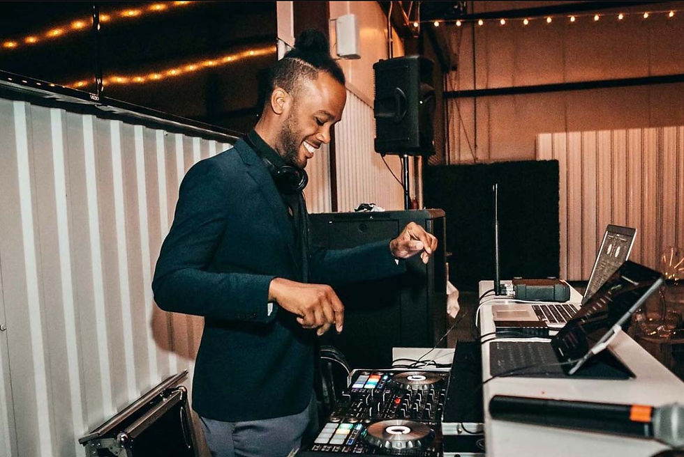 Smiling DJ in a dark blazer mixing music on a controller beside laptops under warm string lights indoors