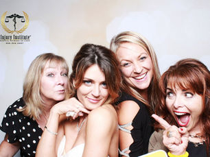 Four women pose and smile in a photo booth, one pointing playfully; Injury Institute logo and phone number appear top left.
