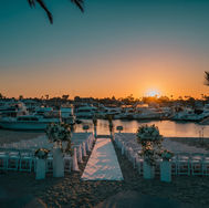Beachside wedding setup at sunset with white chairs, floral arch, and a sandy aisle overlooking yachts on a calm marina