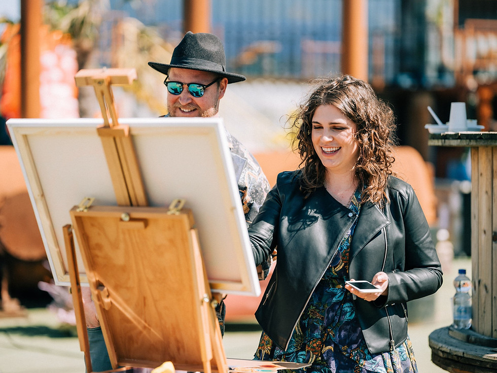 Wedding painters Monica and Villa from Portugal behind easel, laughing, sunny outdoor scene