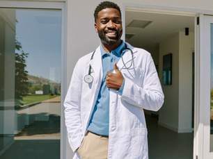 A smiling physician in a white lab coat and stethoscope giving a thumbs up outside a modern medical building