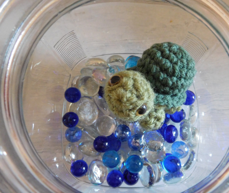A green crochet baby turtle pet in a little jar with blue marbles in the bottom.