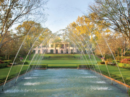 Formal garden with a symmetrical fountain in front, spraying water arcs. Trees and a large mansion are in the background, under a clear sky.