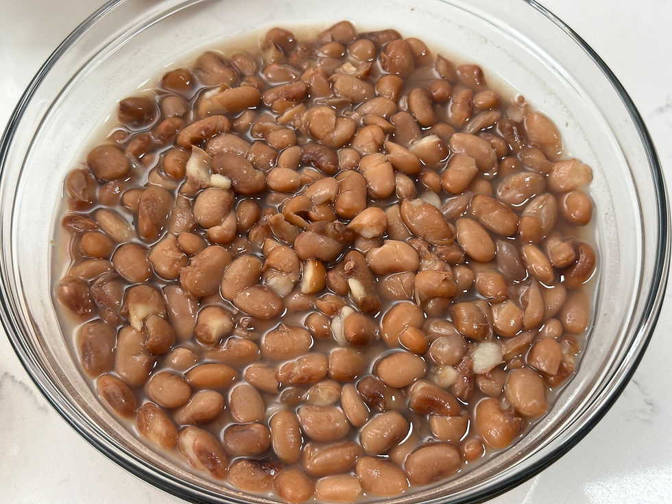 Bowl of cooked pinto beans in liquid on a white surface. The beans are brown and glistening, creating a simple, comforting visual.