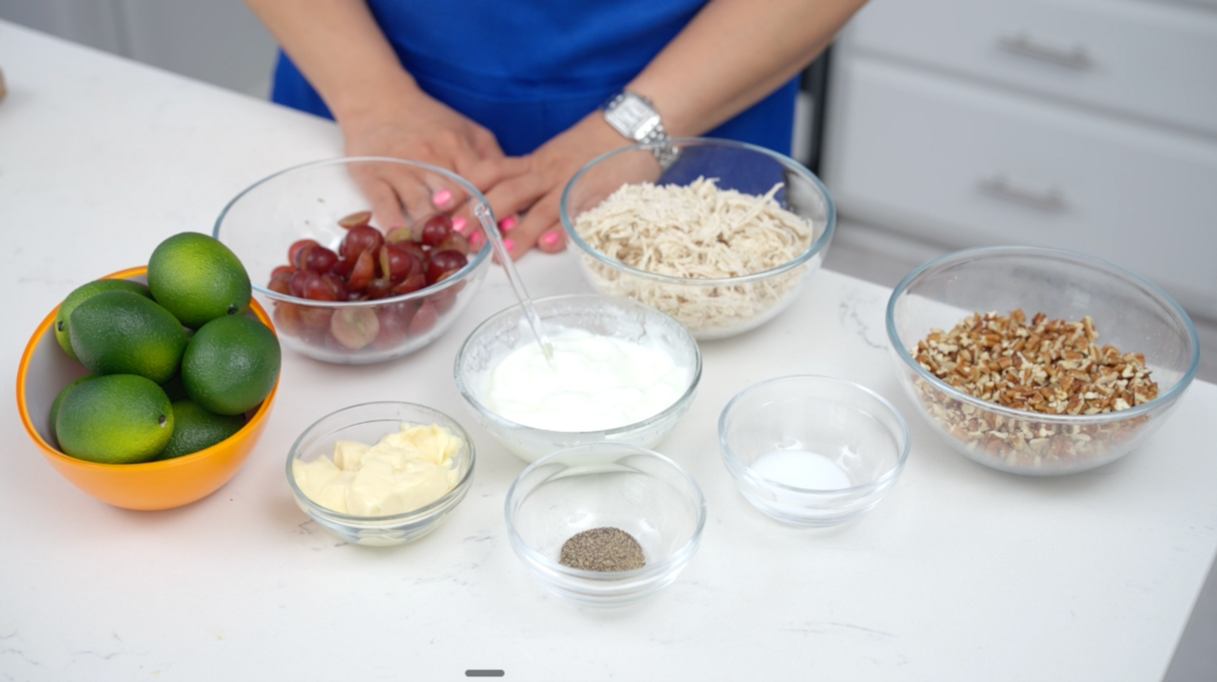 Hands of a person in a blue apron over bowls of mangoes, grapes, coconut, yogurt and nuts on a white kitchen counter.