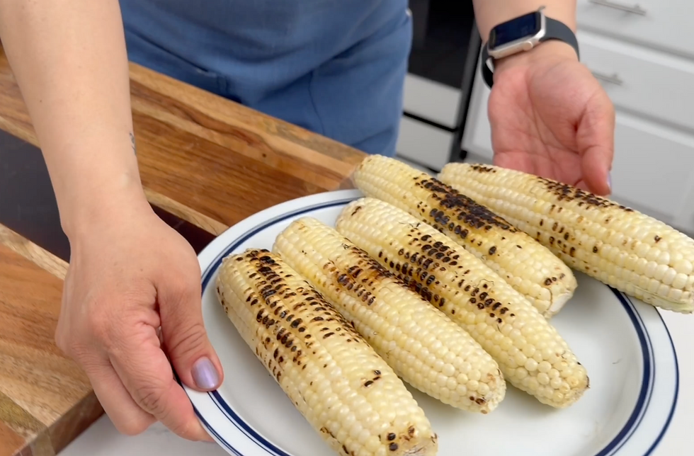 Person holding a plate of grilled corn on the cob in a bright kitchen.