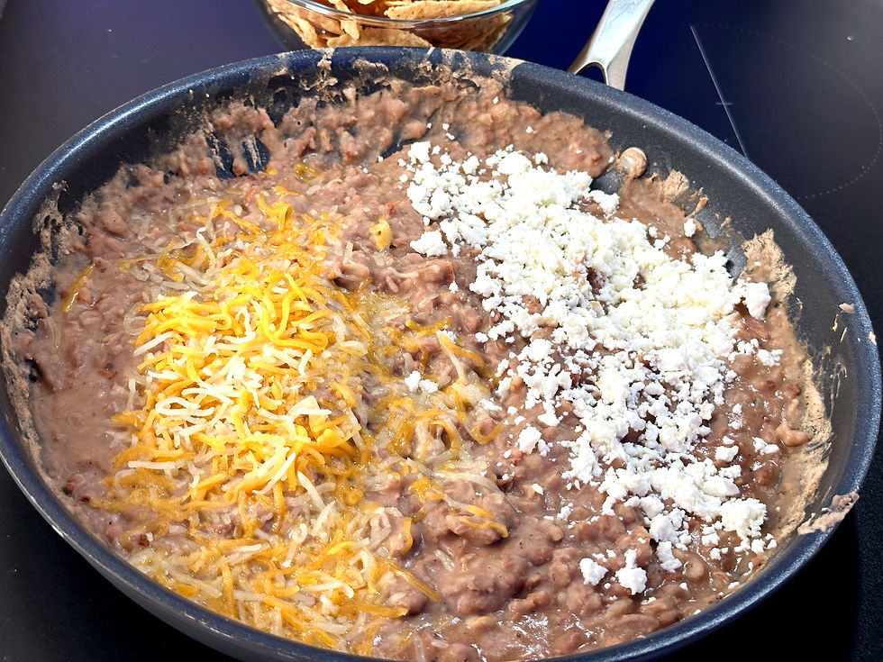 Refried beans with cheddar and crumbled cheese in a pan, over a black stove. A bowl of chips is in the background.