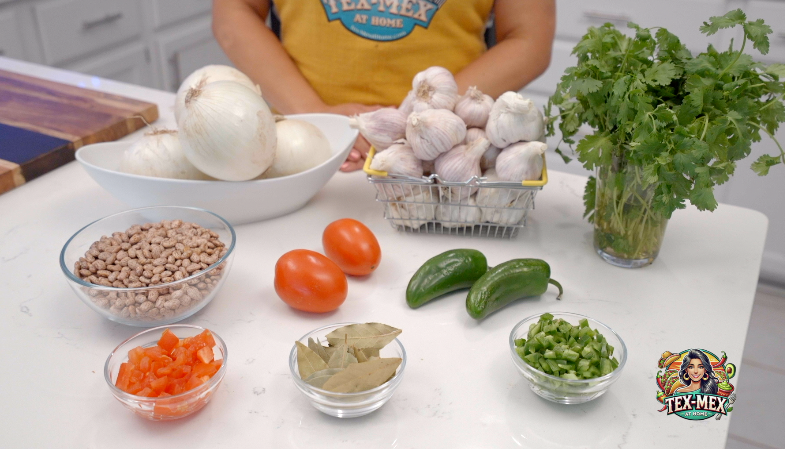 Ingredients on a kitchen counter: onions, garlic, cilantro, tomatoes, jalapeños, beans, and spices. Person in yellow apron with "Tex-Mex at Home" logo.