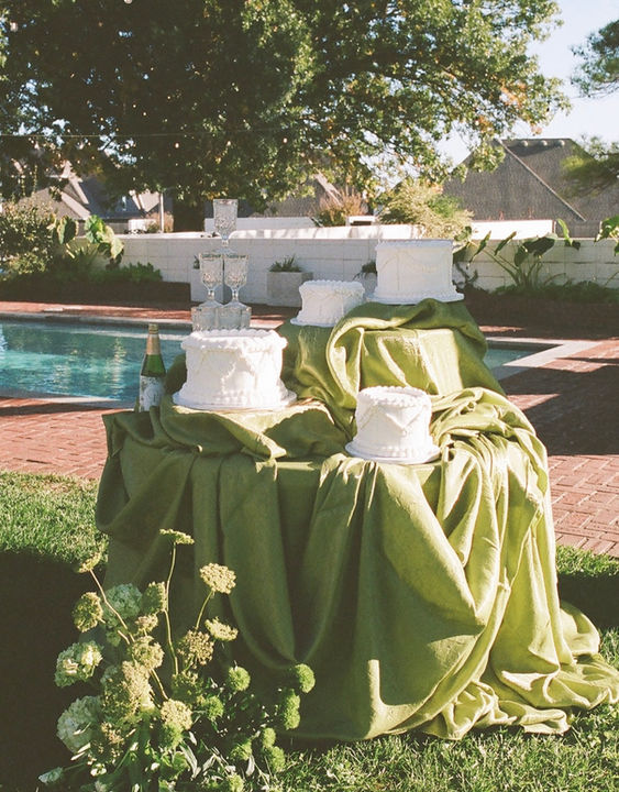 Cake table with green draping table cloths and a floral arrangement sitting by a pool