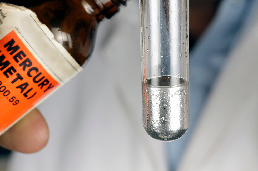Brown bottle labeled "Mercury Metal" pouring into a test tube filled with silver liquid. Background has a blurry figure in a white coat.
