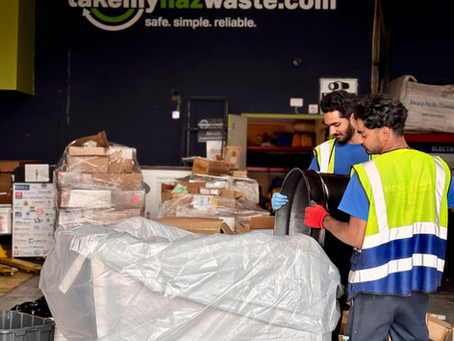 Two workers in yellow vests handle a barrel in the takemyhazwaste warehouse with stacked boxes. They are processing hazardous waste for destruction. Background sign reads "takemyhazwaste.com." Mood is busy.