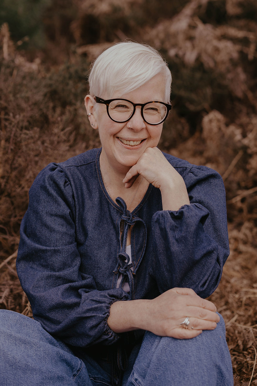 A person with short white hair and glasses smiles warmly, seated in nature. They wear a blue outfit, radiating a relaxed, joyful mood.